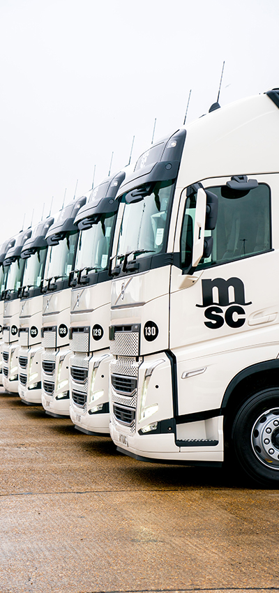 Fleet of MSC trucks parked in a row. The trucks are branded with the MSC logo and are set against a backdrop of a depot. The scene conveys a sense of organised logistics and transportation efficiency.