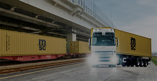 Truck transporting yellow MSC containers on a road beneath an overpass.